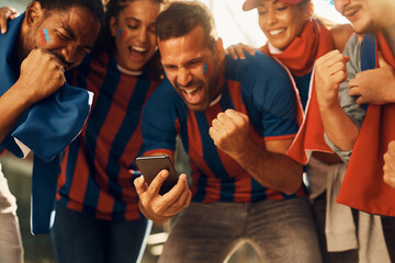 Excited soccer fans celebrating winning goal of their favorite team while watching match on smart...