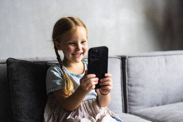 Portrait of cute little cute girl holding smartphone and resting at sofa at home. Choosing favorite music or cartoons,texting messages, browsing internet, watching video, playing games onphone