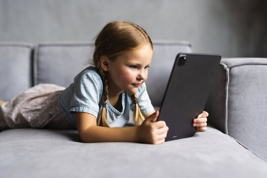 My favorite pastime. Smiling small child girl sit on comfy sofa in modern studio apartment hold tablet computer read book online use handy web app. Little kid rest on cozy couch play videogames on pad