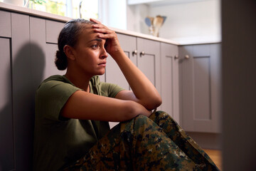 Depressed Female Soldier In Uniform Suffering With PTSD Sitting On Kitchen Floor On Home Leave