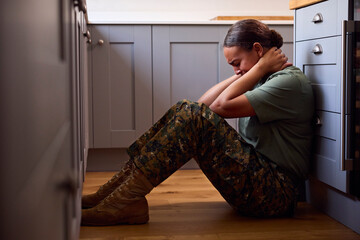 Depressed Female Soldier In Uniform Suffering With PTSD Sitting On Kitchen Floor On Home Leave