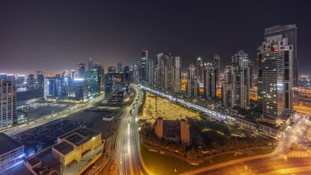 Bay Avenue with illuminated modern towers residential development in Business Bay aerial timelapse panorama during all night with moon, Dubai, UAE. Skyscrapers with traffic on a road near big parking