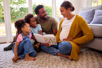 Army Family With Pregnant Mother Sitting On Floor In Lounge At Home Reading Book Together