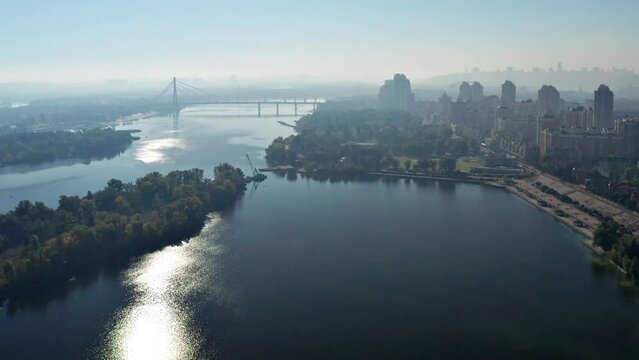 A beautiful flight over Kyiv and the Dnieper River. The sun is reflected in the river. Fog and bridge across the river in the background. Multi-storey buildings along the embankment of the Dnieper.