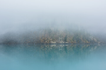 Fototapeta premium Simple meditative landscape with turquoise lake in thick fog. Tranquil view through clearance in dense fog to steep slope in fading autumn colors and mirror mountain lake. Reflective azure alpine lake