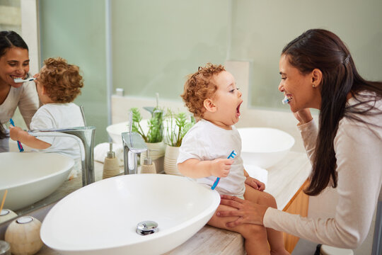Mom teaching baby to brush its teeth, on the bathroom counter in home and a clean smile on her face. Healthy oral hygiene for kid means using child friendly toothpaste, toothbrush and dental routine