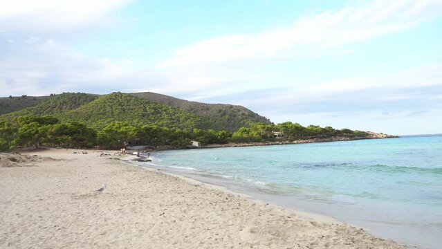 The waves splashing on beach coastline of Cala Agulla (Cala Ratjada) on Mallorca in the mediterranean sea. Deep blue water and a green pine forest in the background. People bathing,  4k.