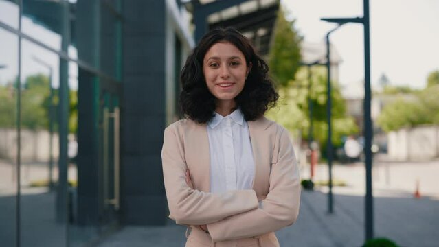 Portrait of the beautiful curly smiling young businesswoman in formal outfit standing near the business centre crossing her hands looking at the camera. People and business concept