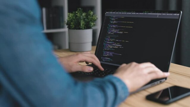 Man in denim shirt coding software at laptop, desk with computer writing code, office workplace