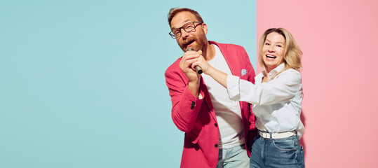 Funny couple of young happy man and woman singing at microphone isolated over blue and pink background.