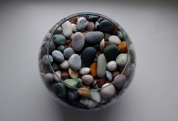 Round glass vase with colorful smooth pebbles and shells on a white surface isolated top view
