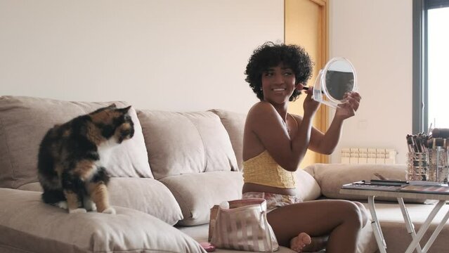 Transgender woman looking at cat and smiling while doing her makeup sitting on sofa at home.
