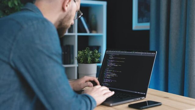 Bearded coder man in denim shirt writing code using laptop, focused male coding in dark office