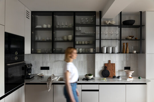 Modern composition of dining space with design kitchen island, furnitures, kitchen stuff, cup of coffee and elegant personal accessories. Woman walking through space. Stylish home decor. Template.  