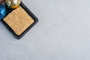 Piece of cake and christmas baubles on a small, black tray on marble background