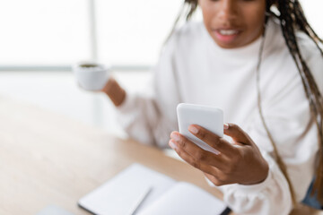 Cropped view of blurred african american businesswoman using smartphone and holding cup in office.