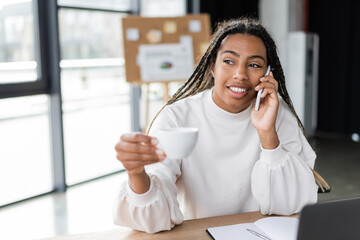 Smiling african american businesswoman holding cup and talking on smartphone in office.