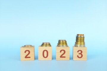 Year 2023 economy growth and recovery, save, invest and grow money concept. Wooden blocks 2023 with increasing stack of coins on blue background.