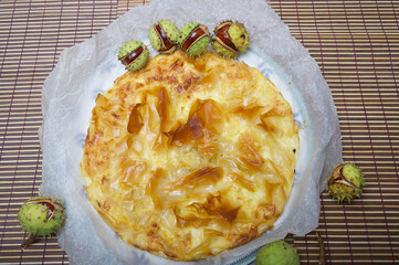 Freshly cooked hot cheese pie in a round plate in a home kitchen - top view. Around the dish lie green fallen chestnuts to create an autumnal romantic mood.