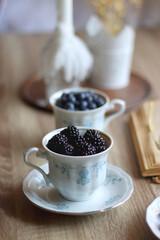 Fototapeta Vintage tea set filled with blueberries, blackberries and starwberries, open book, reading glasses and decorative candles on the table. Selective focus. obraz