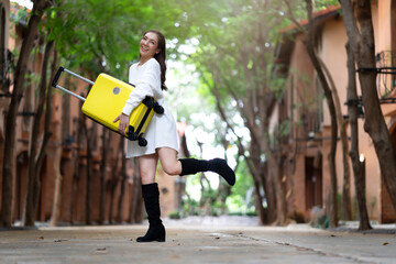 Young cheerful woman traveler with white dress and long black boots holding yellow suitcase in hands, happiness smiling with outdoor natural green background, excited happy girl tourist with luggage
