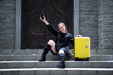 Young beautiful woman sit on stair outside the accommodation with yellow luggage and phone in hand, waiting for her friend to pick her up, showing her hand up to say Hello when seeing friend arrive