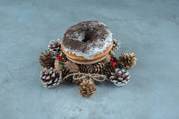 A glazed donut nested on a small wreath amid pine cones on marble background