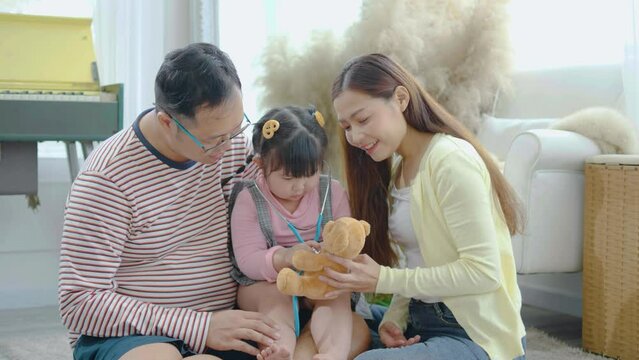 Asian little girl plays with teddy bear toy listens to him with a stethoscope with father and mother, listening to teddy bear patient heartbeat , enjoying game