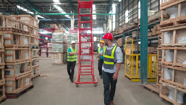 Caucasian male engineer guides two male workers is draging red ladder. Employees wear safety hat and reflective vests working in a warehouse, team work, helping each other.
