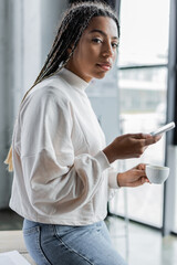 African american businesswoman in sweatshirt holding coffee and smartphone in office.