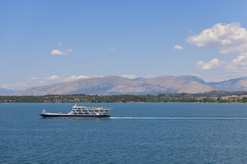 Local small blue and white passenger ferry crosses Garitsa bay against the backdrop of the central Corfu island's high hills with forested coastline, Ionian islands, Greece