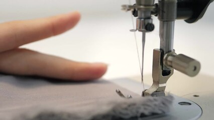 Close up slow video of woman's hand sews grey fabric on a professional sewing machine at the workplace. The seamstress's hands hold textiles for the production of dresses. Light blurred background.
