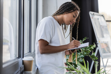 African american businesswoman writing on notebook near blurred coffee to go on windowsill in office.