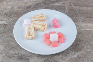 Marmelades, marshmallows and cake roll slices on a platter on marble background