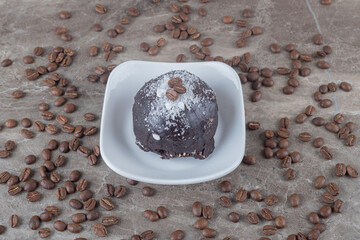 Scattered coffee beans around a small platter with a chocolate coated cake on marble background