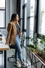 Side view of african american businesswoman in jacket and jeans looking at window in office.
