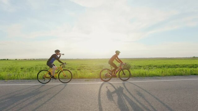 Caucasian men in helmets and sportswear riding bicycles along highway. One sporty cyclist drinking water from bottle with summer sunset on background.