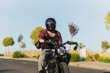 Chico joven tatuado con camisa a cuadros roja montando motocicleta por carretera solitaria al atardecer