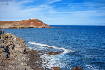 A view of the ocean and a small lighthouse at the entrance pier of the island. Calm blue sea and sky, stone shore.