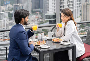 Young couple enjoy breakfast table outdoor on rooftop skyscraper building in cityscape background during sunlight in the morning. Diverse ethnic couple sitting together on roof balcony in skyline view