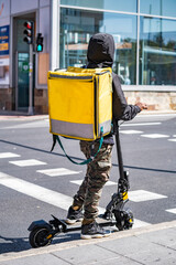 Man courier online food delivery with yellow thermal backpack waiting the green traffic light on an electric scooter.
