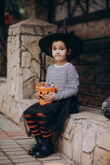 Little cute girl in witch costume holding jack-o-lantern pumpkin bucket with candies and sweets. Kid trick or treating in Halloween holiday.