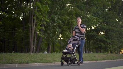 Obraz premium A man rolls a baby around the park and eats a burger. Young father with his son for a walk.