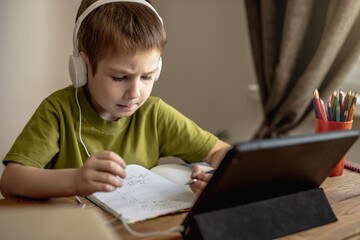 The child is a boy in a green T-shirt with headphones doing homework on a tablet. Concept of remote school education