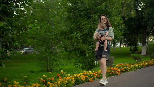 A young woman walks along the alley in the park with her son. The girl in glasses walks. Newborn son in a kangaroo-backpack.