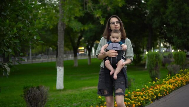 A young mother walks in the park with a child in her arms. Woman with glasses with her son. Newborn baby in a kangaroo-backpack.