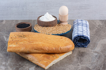 Egg cup, flour bowl and wheat pile on a platter next to black sesame bowl, roll of towel, and bread loaves on marble background