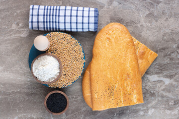 Egg cup, flour bowl and wheat pile on a platter next to black sesame bowl, roll of towel, and bread loaves on marble background