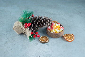 Decoration piece made of pine cone next to dried lemon slices and a bowl of candies on marble background