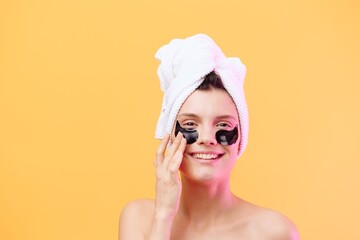 Horizontal photo, a woman with perfect skin on an orange background in a towel on her head and body with black patches on her face looks away and plays with her hands